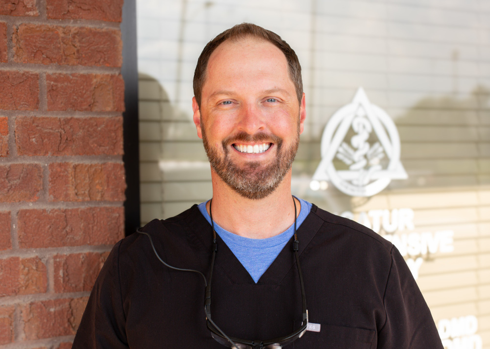 Dr. Brian G. Ondocsin, DMD, in black scrubs with dental loupes around his neck, smiling outside the practice in Decatur, Alabama